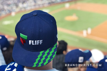 Man wearing a dark blue baseball cap with Fliff logo and green money pattern, overlooking a baseball stadium. 