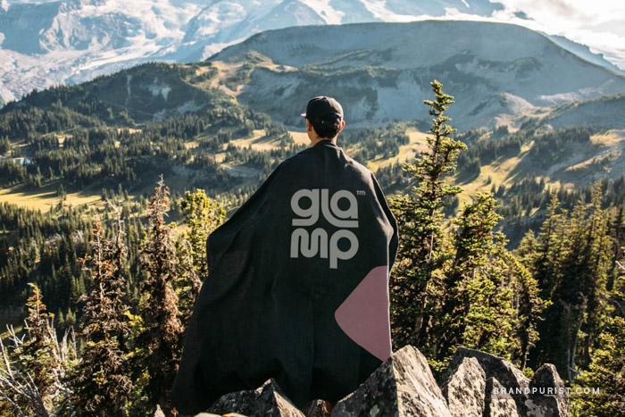 Photo of a young person, seen from behind, draped in a Glamp branded blanket, standing on a hill and gazing towards a distant range of mountains.