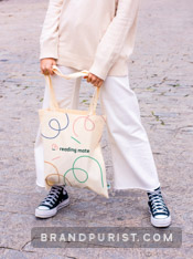 Young person dressed in white, holding a canvas tote bag with Reading Mate’s branding.